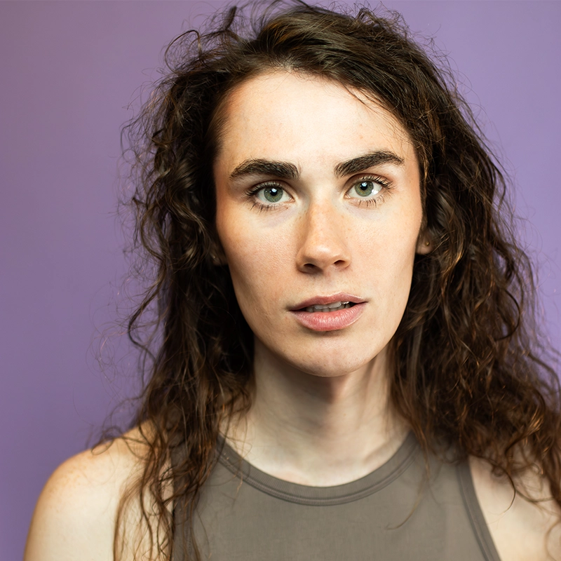 A person with long, curly brown hair and green eyes looks directly at the camera. They have light skin and wear a sleeveless, sheer gray top. The background is solid purple, creating a strong contrast with their features.