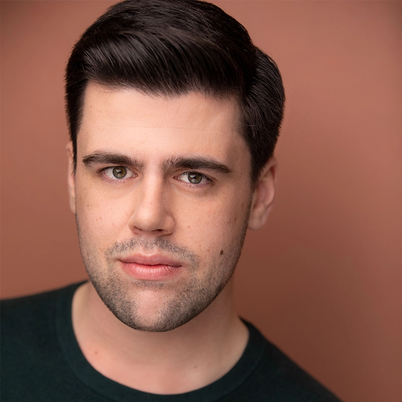 A young man with neatly styled dark hair and light stubble looks directly at the camera. He wears a dark green sweater and stands against a solid, warm brown background, with a neutral expression on his face.