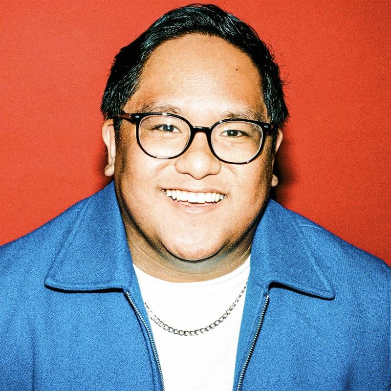 Raymond Salgado, with short dark hair, wearing glasses, a bright blue jacket, a white shirt, and a silver chain necklace, is smiling broadly against a vibrant red background.
