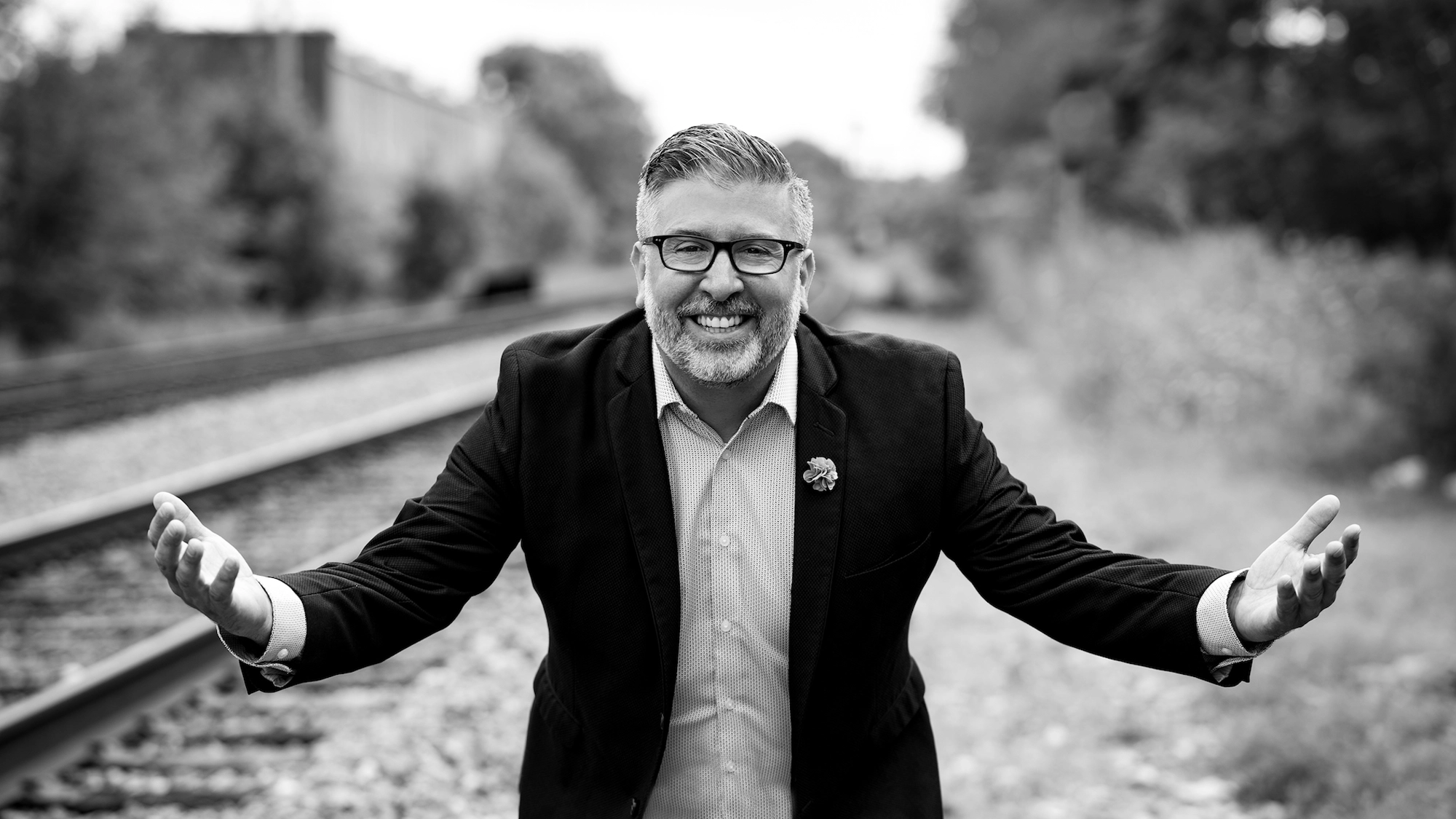 A man with glasses and a beard, dressed in a suit jacket and shirt, stands on railroad tracks with his arms open and smiling broadly—channeling Broadway charm. The black-and-white image features Sam Fazio with blurred trees and tracks in the background.