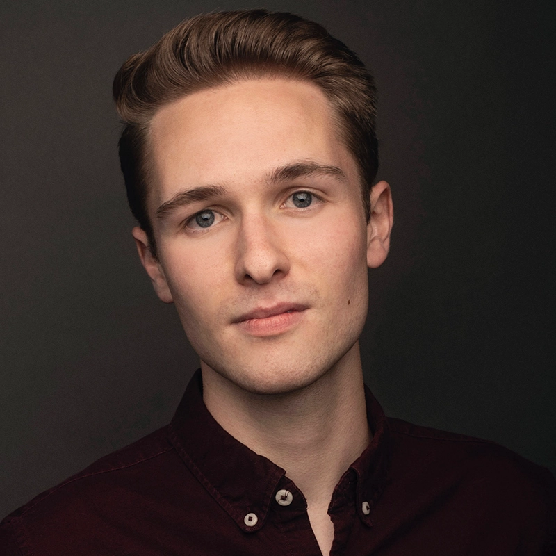 A young man with fair skin and neatly styled light brown hair poses against a dark background. He wears a burgundy button-up shirt and looks directly at the camera with a calm, neutral expression. The lighting is soft, highlighting his facial features.