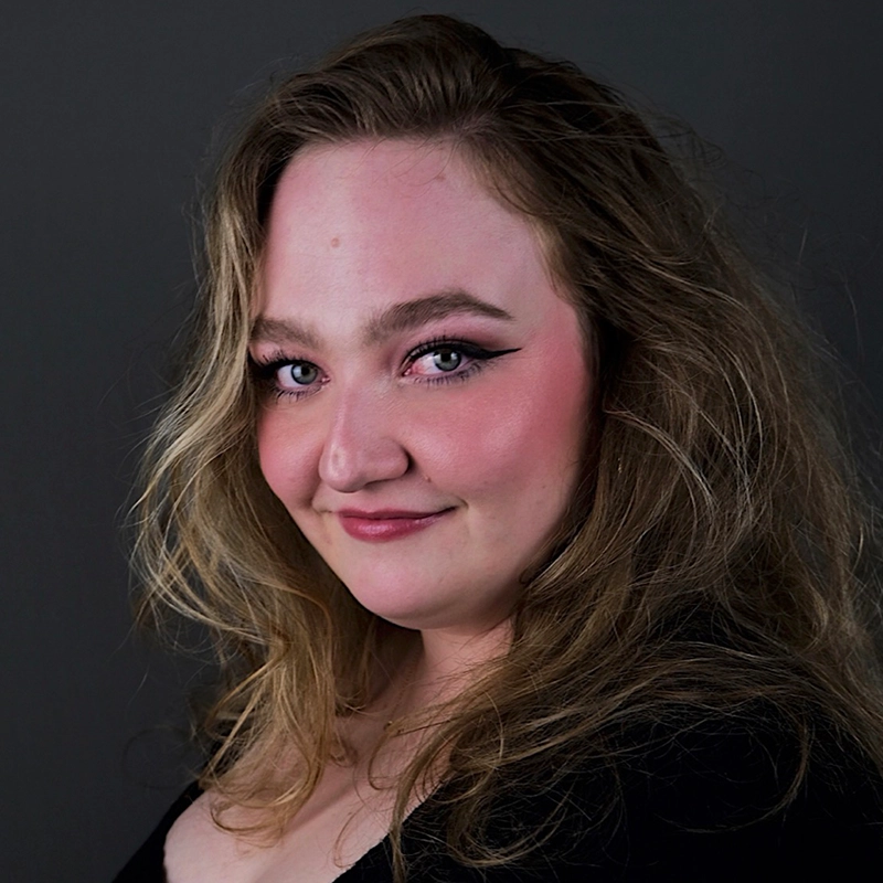 A woman with wavy, light brown hair and fair skin poses against a dark background. Jett Roberts wears pink blush, bold eyeliner, and pink lipstick with a black top, smiling slightly and gazing confidently at the camera.