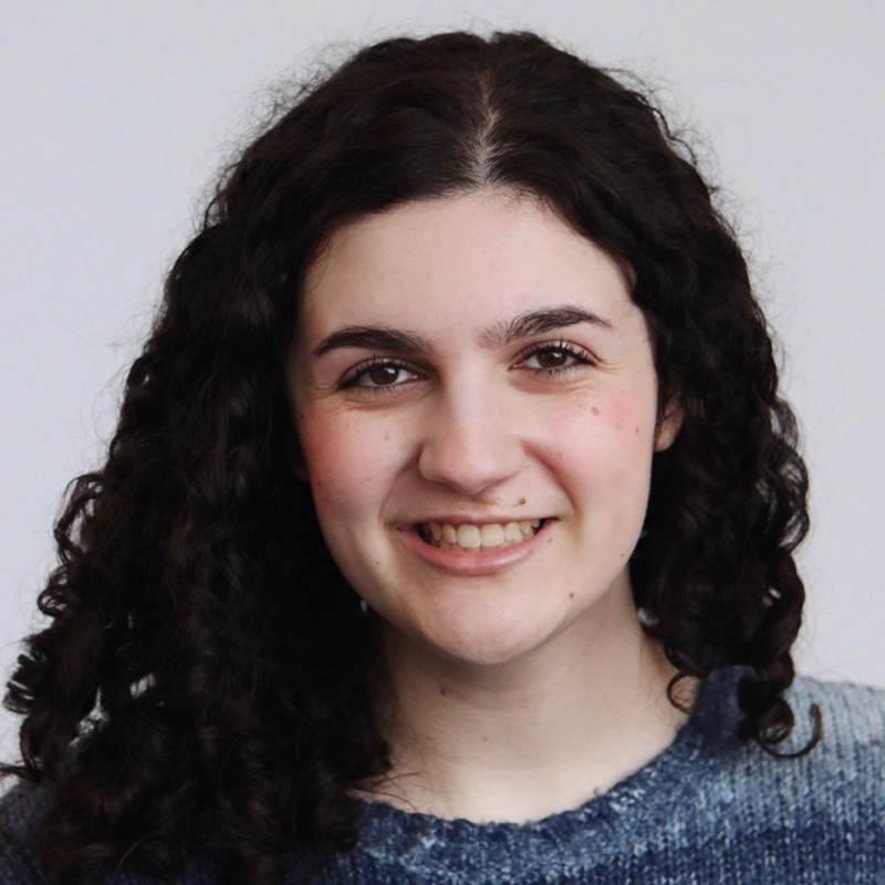 A young woman with long, dark curly hair smiles at the camera. She is wearing a blue, textured sweater and is posed against a plain, light grey background. Her expression is warm and friendly.