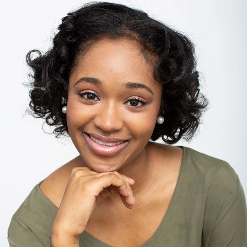 A young woman with short, curly black hair and pearl earrings smiles warmly. She is wearing an olive green top and resting her chin on her hand. The background is plain and white, creating a bright, friendly portrait.