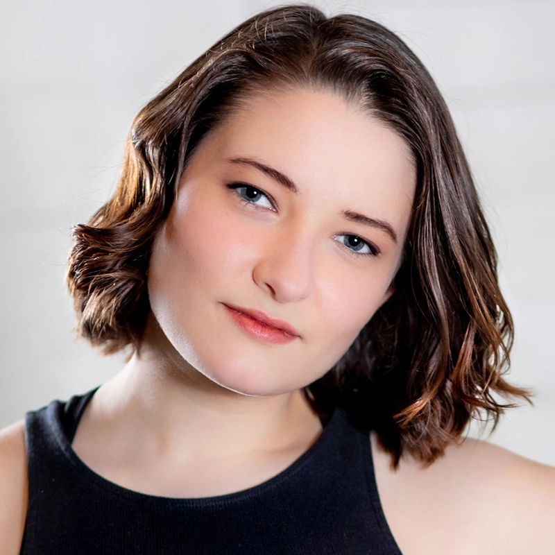 Gabriella Santalla, a woman with fair skin and short, wavy brown hair parted on the side, looks at the camera with a neutral expression. She wears a black sleeveless top and subtle makeup against a softly blurred light-toned background.