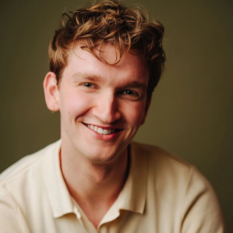 A young person with light skin and wavy, light brown hair smiles warmly at the camera. John Sheldon wears a cream-colored collared shirt and a small hoop earring in his left ear. The background is plain and softly lit in a muted green tone.