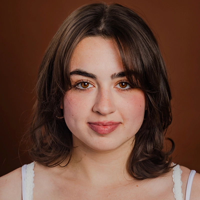Sophia Davison, a young woman with shoulder-length brown hair and brown eyes, looks at the camera in a white top. She has light skin, natural makeup, and a neutral expression against a solid, warm brown background.