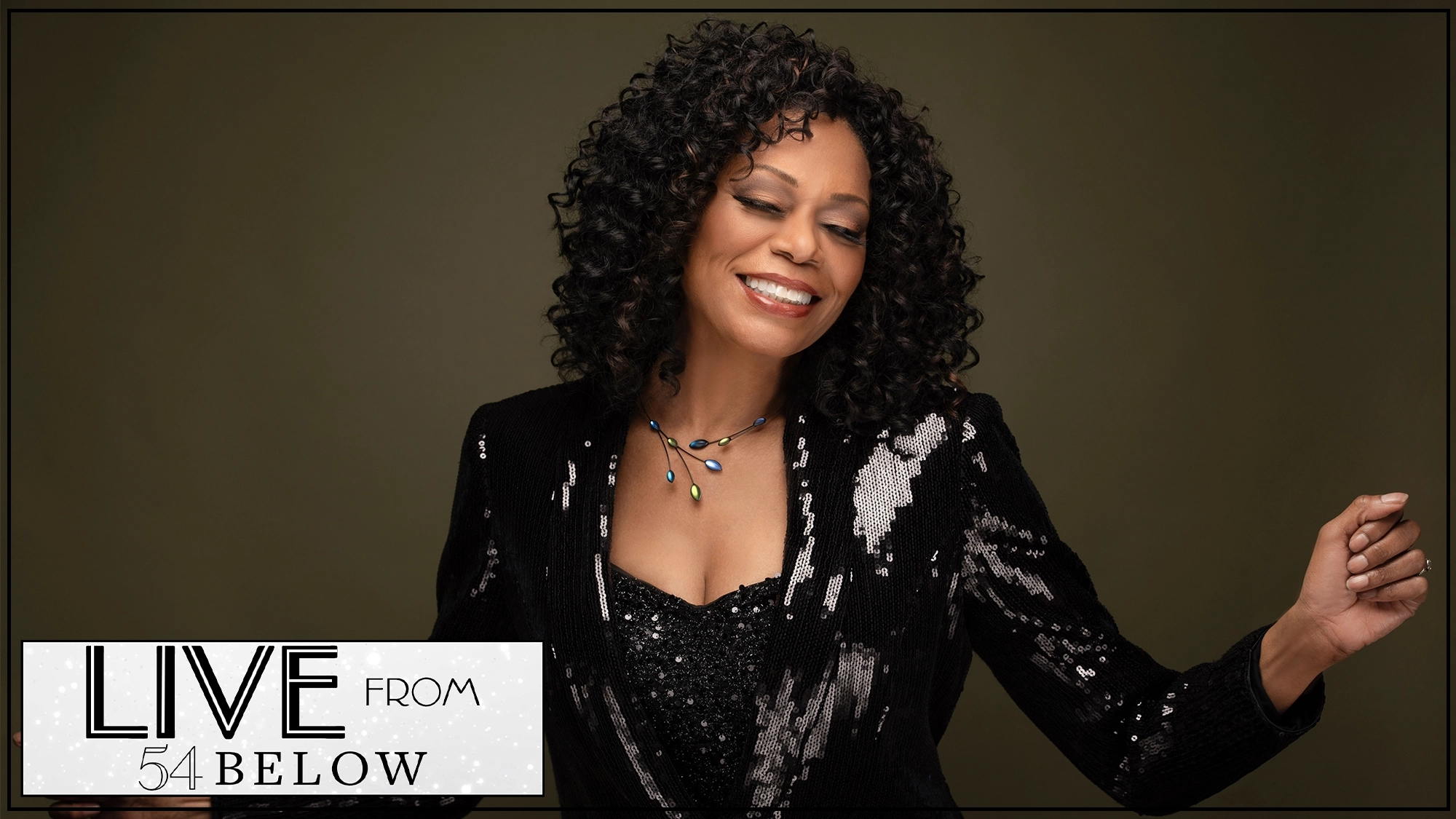 A woman with curly black hair smiles with her eyes closed, wearing a sparkling black outfit and multicolor necklace, posed against a dark background. White text at the bottom left reads “LIVE FROM 54 BELOW.” Livestream starring Stephanie Pope: Livin’ It Up.