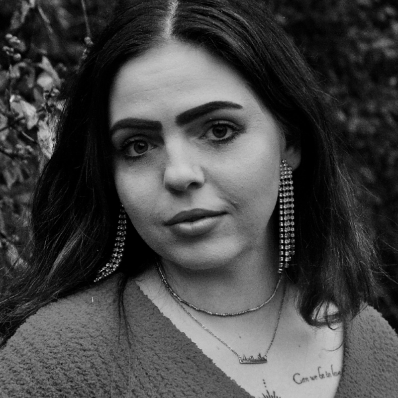 Black and white photo of a woman with long dark hair, thick eyebrows, and large dangling earrings. She wears layered necklaces and a textured top. Some tattoo text is visible on her chest. She stands outdoors with leafy foliage blurred in the background.