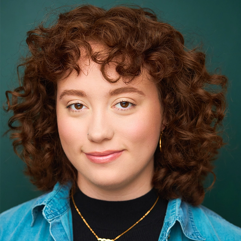 Sydney Kenton, a young person with short, curly brown hair, smiles slightly at the camera. They wear a light denim shirt over a black top, gold hoop earrings, and a gold necklace. The background is solid green; their makeup is natural and subtle.