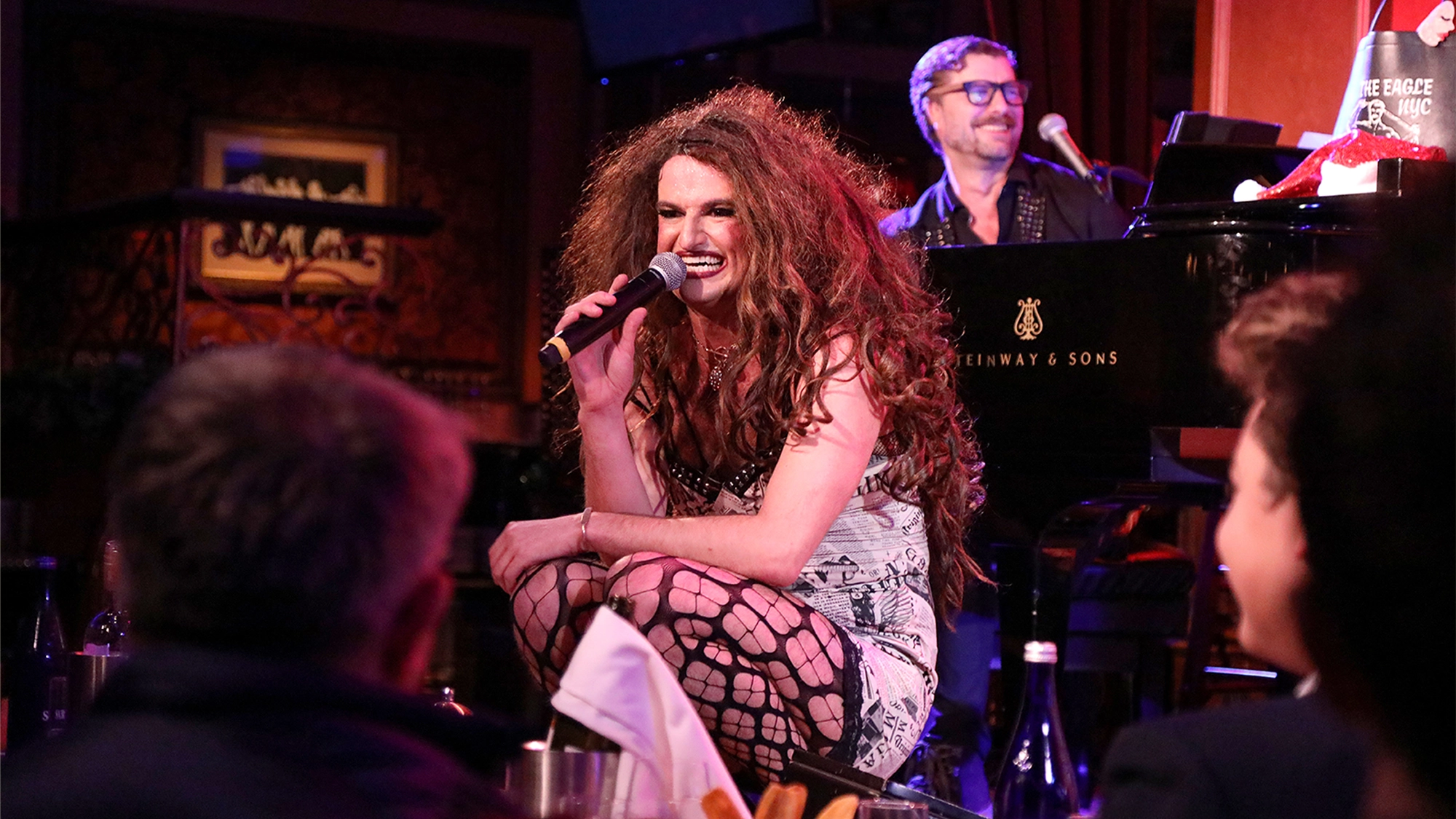 Tammy Faymous site image 26 A performer in a dress and fishnet stockings, part of the Tammy Faymous Experience, kneels and sings into a microphone on stage. A smiling pianist plays a grand piano behind them as the audience enjoys lively entertainment under colorful stage lighting.