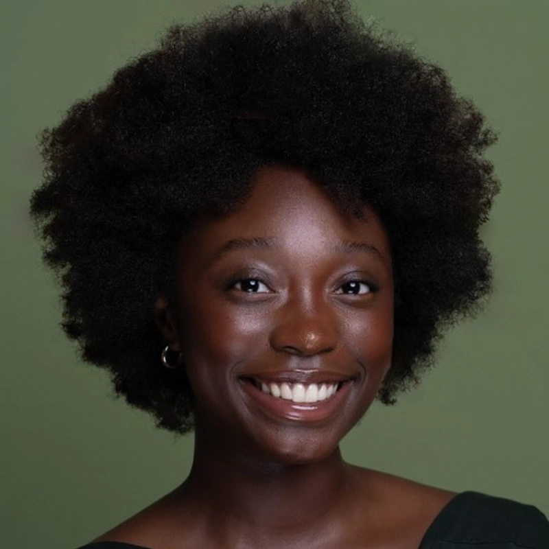 A smiling woman with a natural afro hairstyle, wearing a black top and small hoop earrings, is posed against a solid olive green background. She looks directly at the camera, her clear skin and bright eyes embodying Danté X Johnson’s vibrant style.