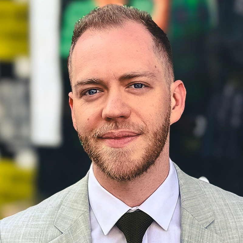 A man with short light brown hair and a trimmed beard, identified as Tom Jorgensen, wears a light gray suit, white shirt, and dark tie. He looks at the camera with a slight smile. The blurred background shows patches of yellow, green, and dark colors.