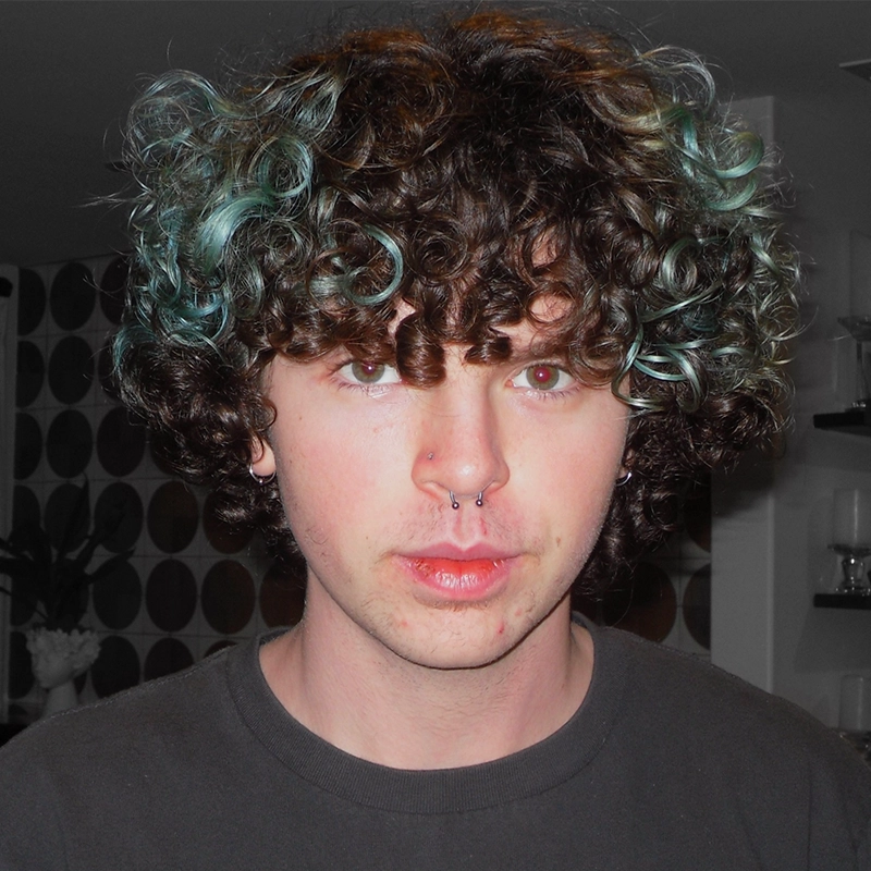 A young person with pale skin, green eyes, a septum piercing, and short curly brown hair with teal highlights, stands indoors against a modern background with circular wall patterns and glass shelves, wearing a dark shirt.