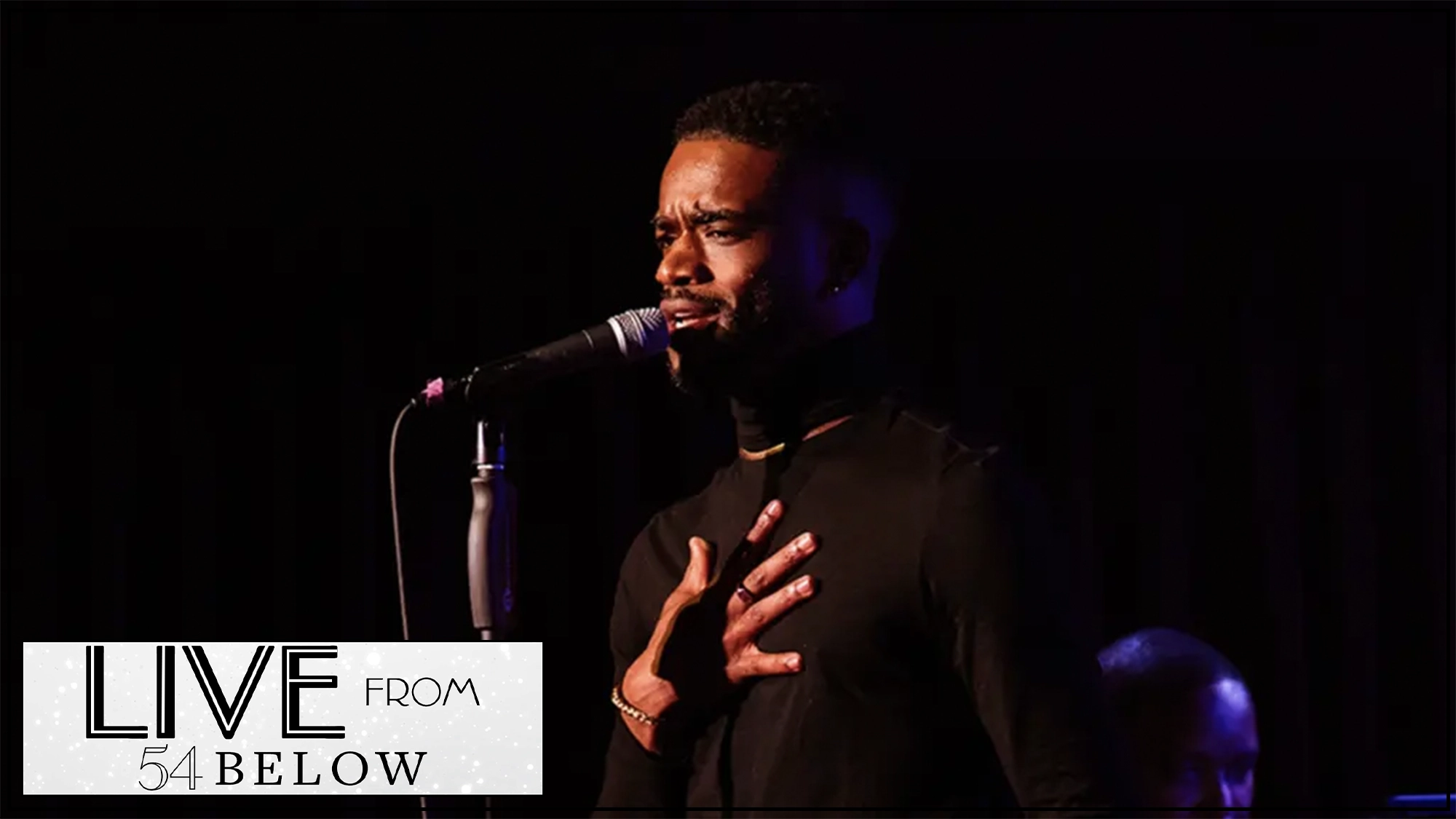 A man in a black turtleneck passionately sings into a microphone on stage, his hand on his chest. The background is dark. In the lower left corner, a “LIVE FROM 54 BELOW” logo appears, featuring Treston J. Henderson's Livestream: The Living Room Session.