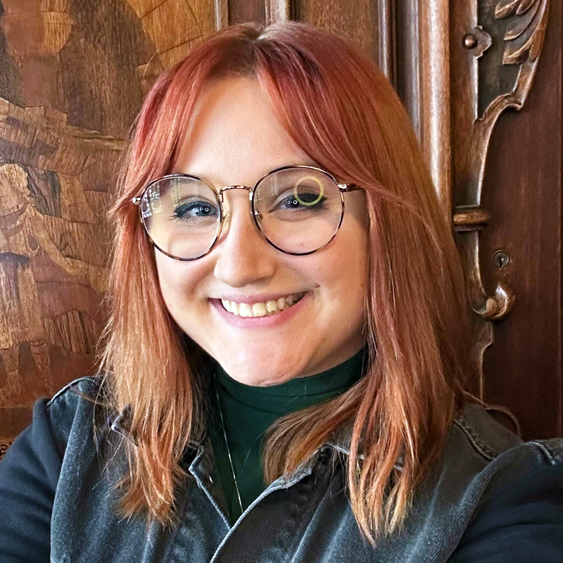 Lucia Troiano, a young woman with shoulder-length reddish-brown hair and round glasses, smiles at the camera. She wears a dark top over a green turtleneck, standing before ornately carved wooden paneling with detailed patterns.
