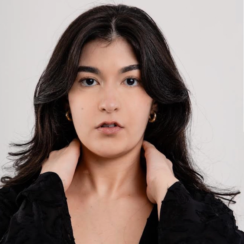 A person with long dark hair and light skin looks directly at the camera, holding their neck with both hands. They wear a black top and small hoop earrings. The background is plain and light-colored.