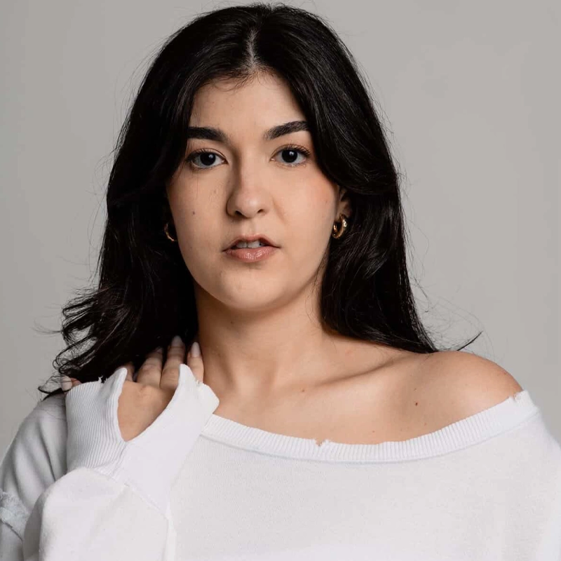 A young woman with long, dark hair and light skin gazes at the camera. She wears a white, off-the-shoulder top with a thumb hole in the sleeve. Her hand gently touches her collarbone. The background is plain and light-colored.