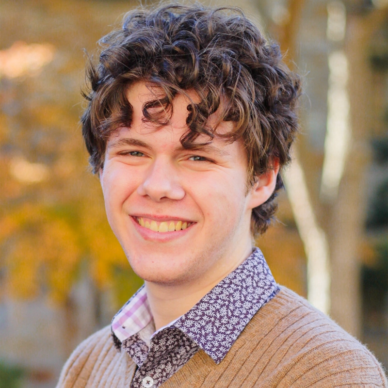 A young man with curly brown hair, Daniel Weintraub, smiles at the camera. He is wearing a tan sweater over a white shirt with a dark floral pattern. The blurred background features autumn-colored trees with golden and orange leaves.