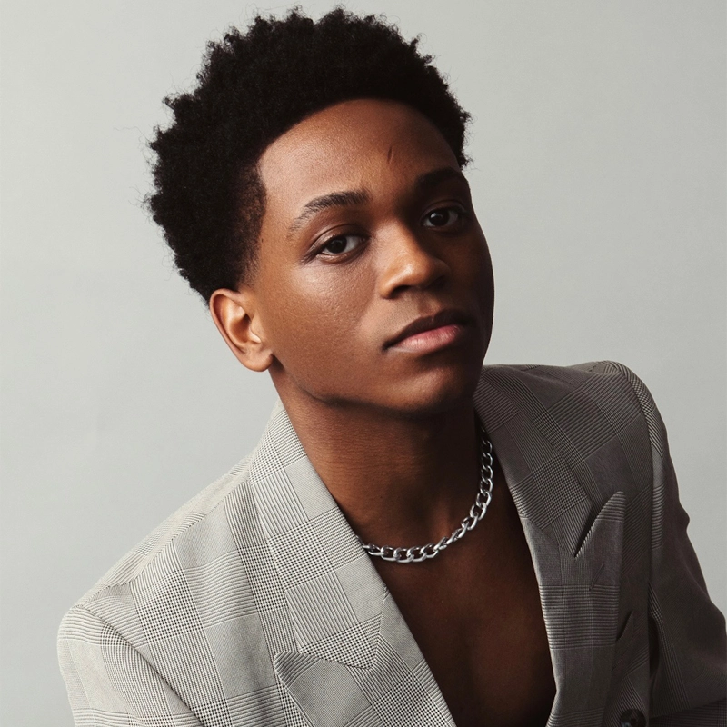 Jayden K. White, a young person with short curly black hair, wears a light gray plaid blazer and a chunky silver chain necklace. They gaze confidently at the camera against a plain light gray background, their expression calm and self-assured.