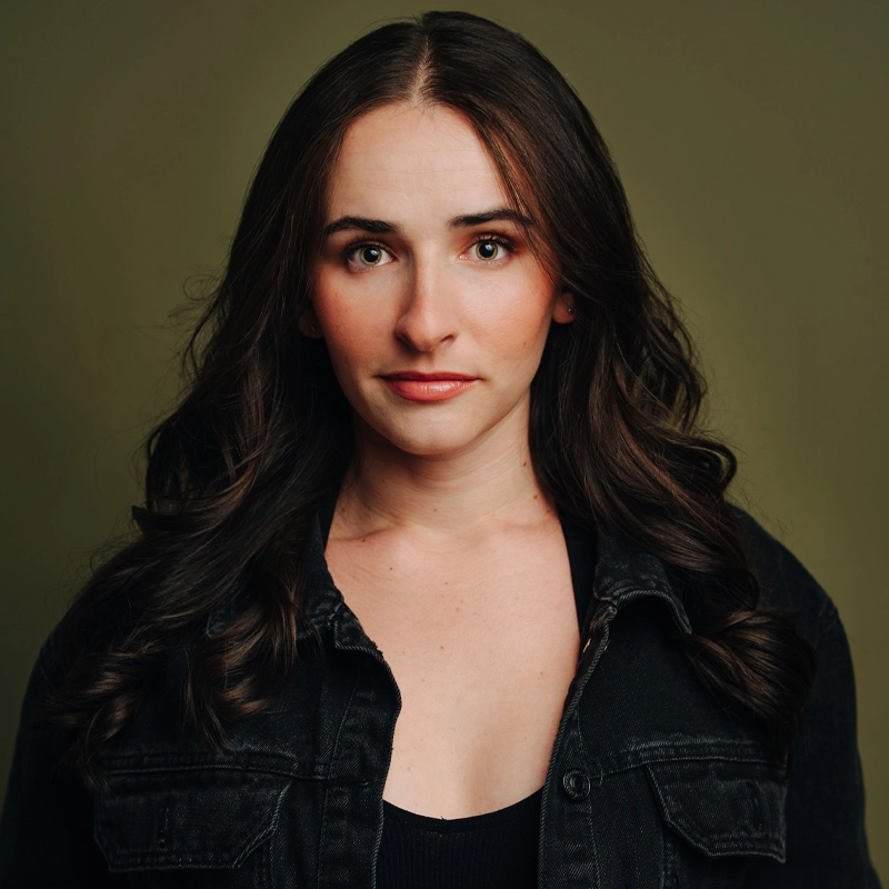 Hannah Wiegand, with long, wavy dark brown hair, stands against an olive green background. She has light skin, subtle makeup, and wears a black denim jacket over a black top, looking directly at the camera with a neutral expression.