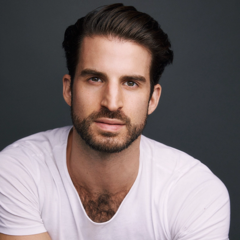 A man with short, slicked-back dark hair and a trimmed beard, resembling Zach Williams, looks at the camera. He is wearing a white t-shirt against a dark grey background, with a neutral expression and soft lighting on his face.