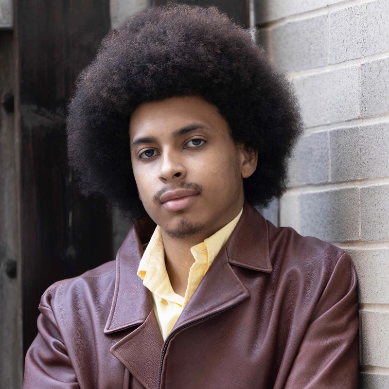 A young man, Jordan Woods, with a large, curly afro hairstyle stands against a brick wall. He is wearing a brown leather jacket over a yellow collared shirt, gazing calmly at the camera with his arms crossed amid wooden and brick textures.