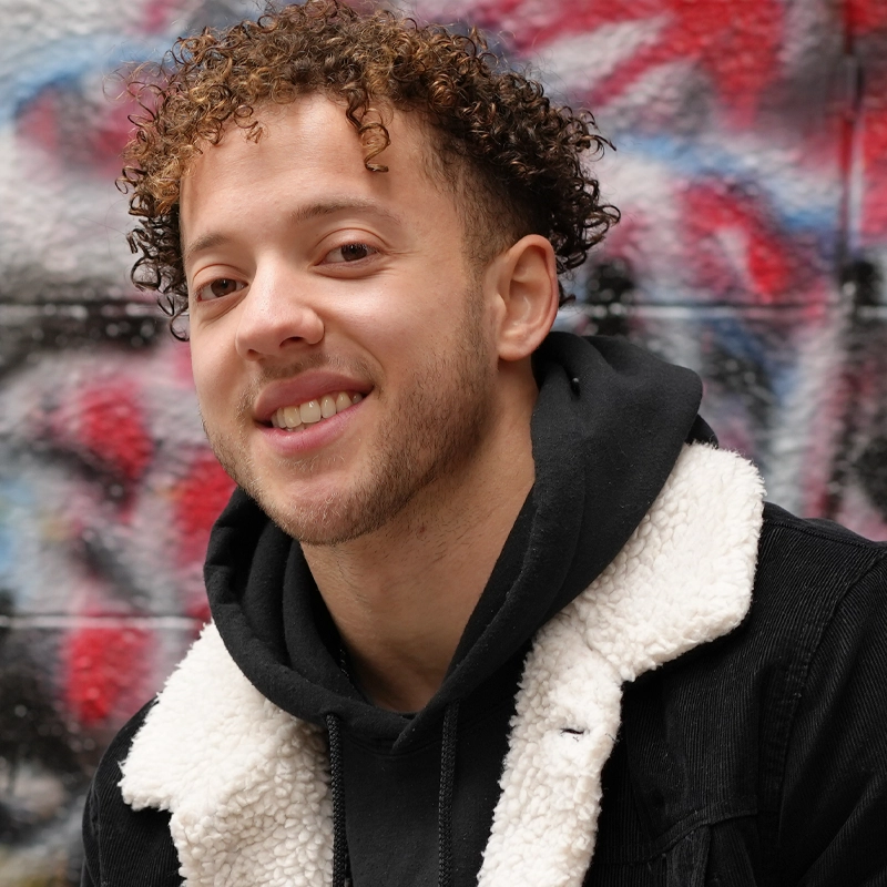 A young man with short curly hair and a trimmed beard smiles at the camera. He is wearing a black jacket with a white sherpa collar and a black hoodie underneath. The background is a wall with colorful graffiti in red, blue, and white tones.