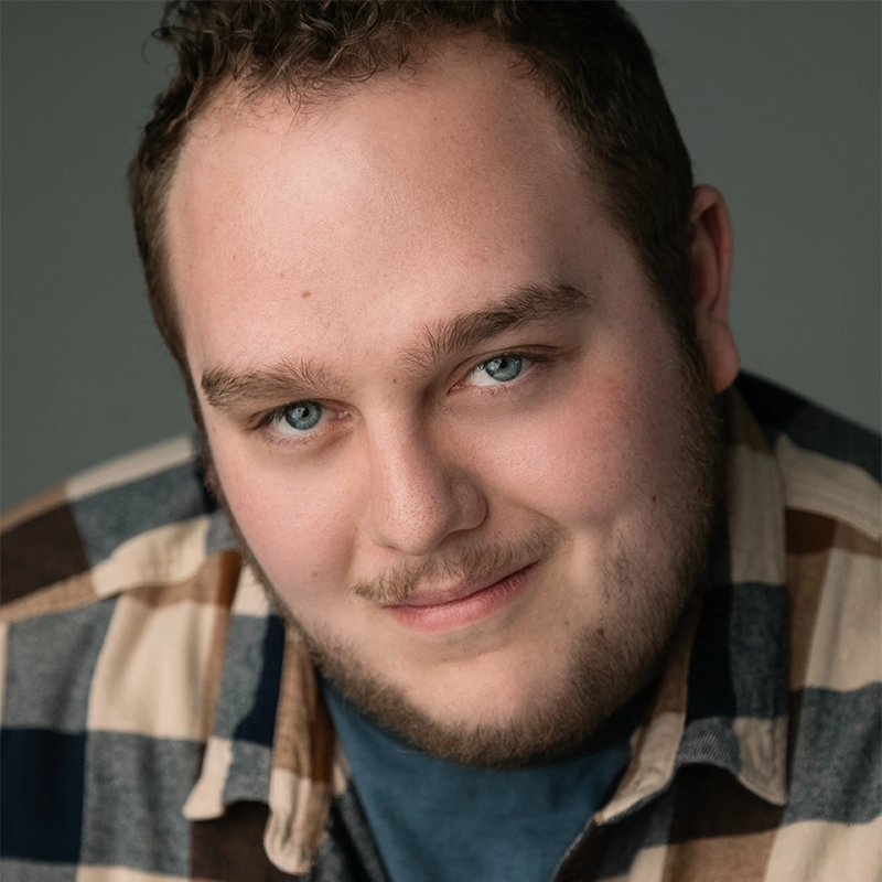 Xavier Wilson, a man with short curly hair, blue eyes, and a trimmed beard, smiles gently. He wears a plaid flannel shirt over a blue t-shirt. The plain gray background and soft lighting highlight his facial features.