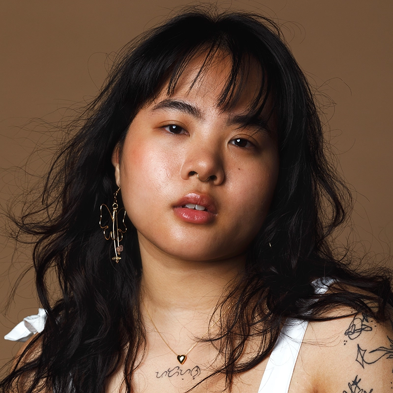 A young woman with long, wavy black hair poses against a brown background. Annie Thomas wears a white top, a gold heart necklace, and an angel-shaped earring. Her expression is neutral, and she has visible tattoos on her arm.