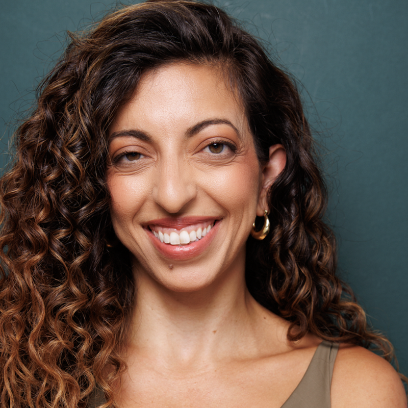 Gabby Ziccarelli, with long, curly brown hair, smiles warmly at the camera. She wears a sleeveless top and gold hoop earrings. Her tan complexion is accented by a beauty mark above her lip, set against a muted green background.
