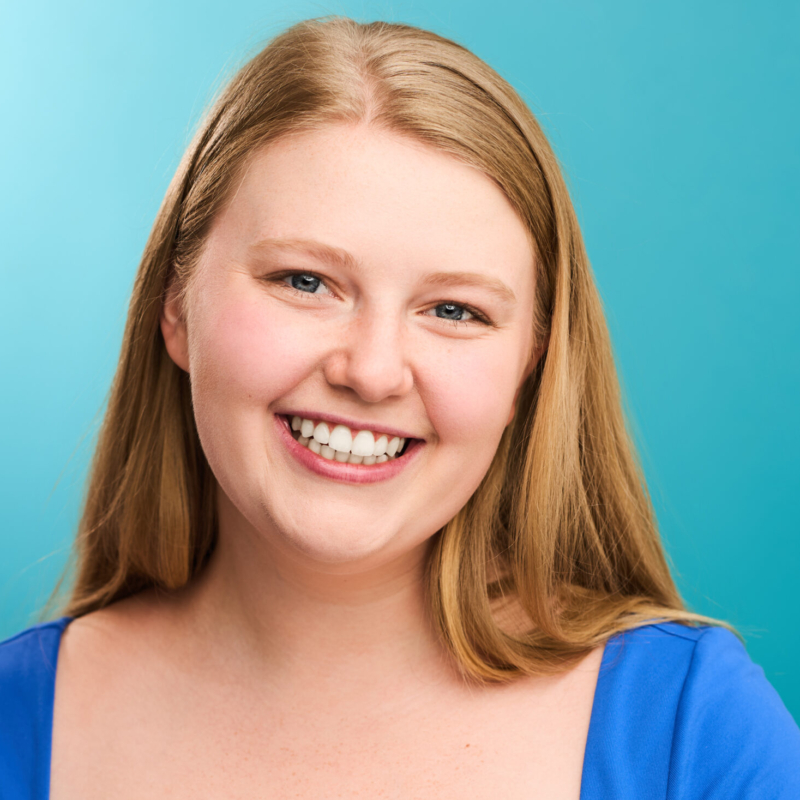 A young woman with straight, long blonde hair smiles broadly. She has light skin, blue eyes, and wears a blue top. The background is a solid light teal color, giving the portrait a bright and cheerful feel.