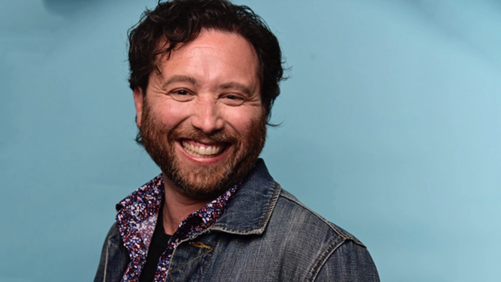 A man with wavy brown hair and a beard smiles broadly at the camera. He is wearing a blue denim jacket over a patterned shirt, channeling Adam Blotner's distinctive style. The background is a solid light blue color.
