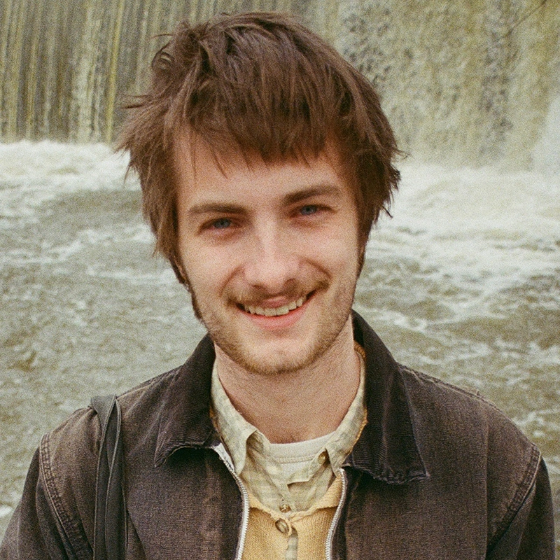 A young man with tousled brown hair and a slight beard smiles warmly at the camera. He wears a dark denim jacket over a button-up shirt. Behind him, water cascades down a wide, blurred waterfall, capturing Maya Monteleone's love of nature.