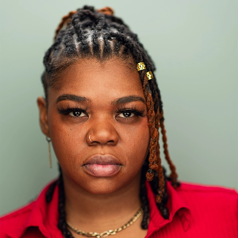 Jenna Leigh Miller, with medium brown skin and styled locs adorned with gold cuffs, looks directly at the camera. She wears a red collared shirt, gold chain necklace, hoop earring, and nose ring against a plain, soft green background.
