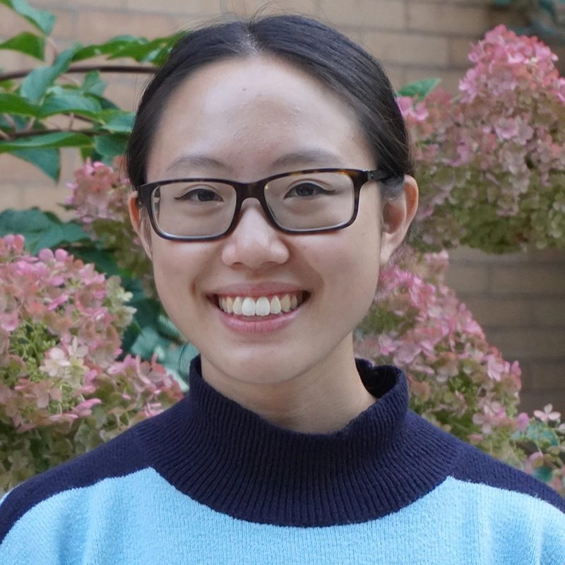 A young woman with dark hair tied back, wearing rectangular glasses and a light blue sweater with a dark collar, smiles in front of pink hydrangea flowers and green leaves against a brick wall background.