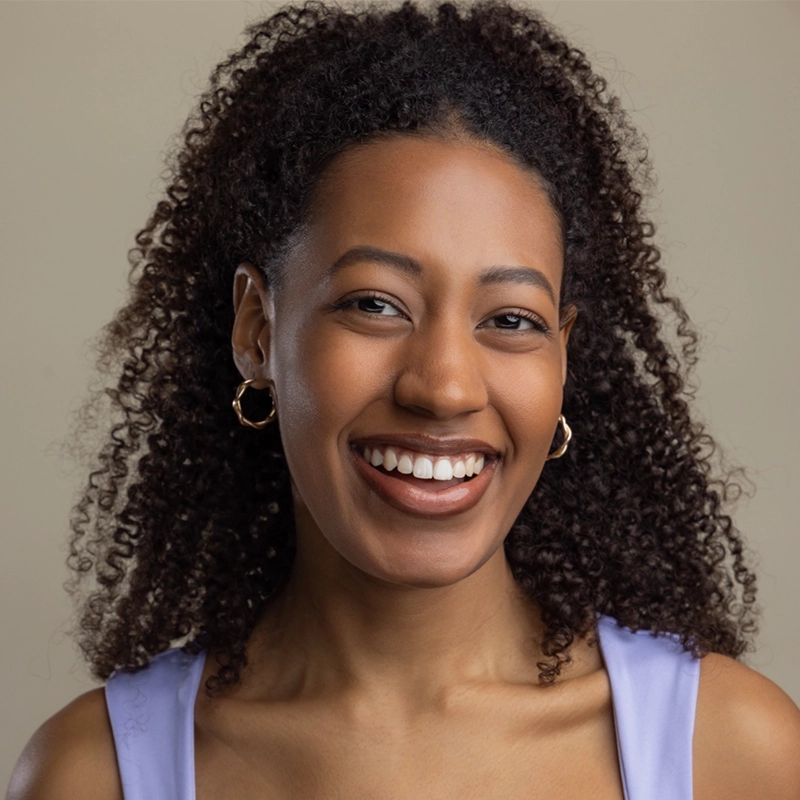 A young woman with curly dark hair smiles warmly at the camera. She wears gold hoop earrings and a light purple sleeveless top. The background is a soft beige, creating a simple and elegant portrait.