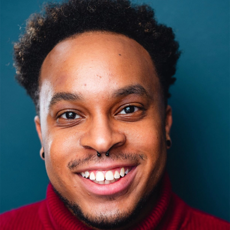 A smiling Chance Anding with short curly hair, wearing a burgundy turtleneck sweater and a septum piercing, stands against a solid teal background. The lighting highlights their cheerful expression and clear skin.