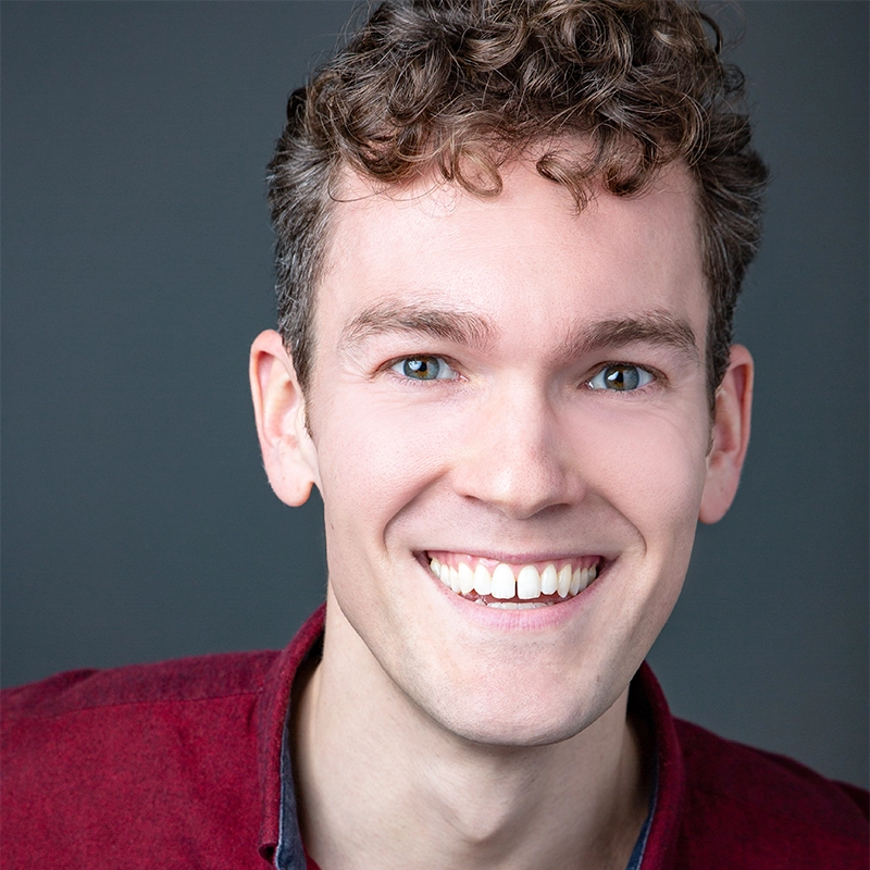 A young man with curly brown hair and blue eyes smiles widely, showing his teeth. Wearing a maroon collared shirt, he stands against a plain dark gray background. The well-lit image highlights his cheerful expression reminiscent of Jenna Leigh Miller’s portraits.