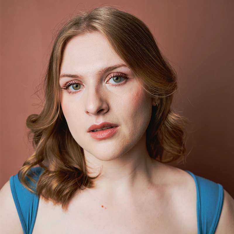 A young woman, resembling Mireya Velasquez, with wavy blonde hair, fair skin, and blue eyes looks directly at the camera. She wears natural makeup and a blue sleeveless top against a soft brown background with gentle, even lighting.