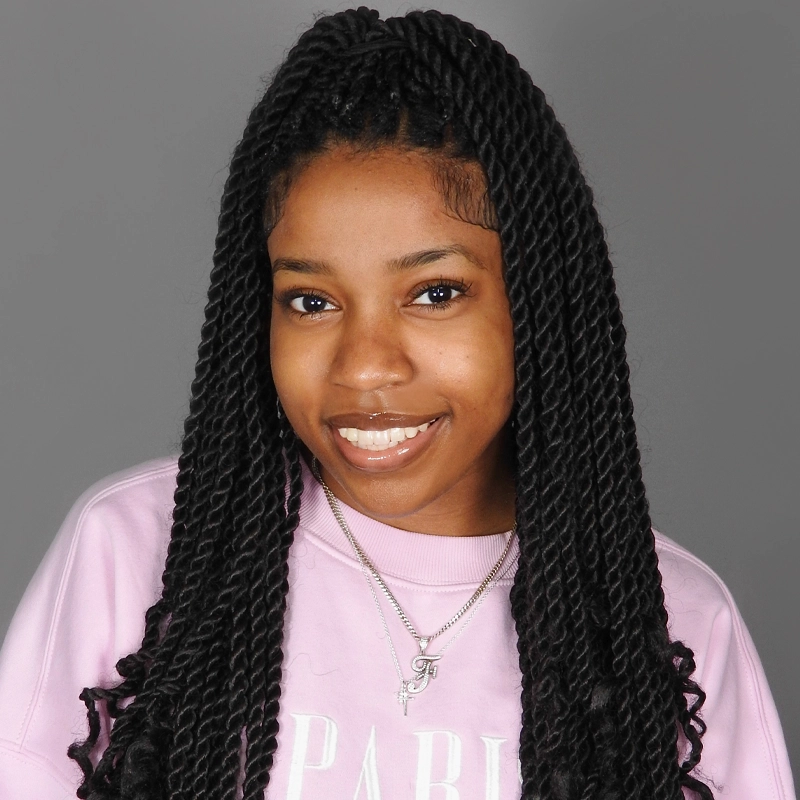 Faith Angeline, a young woman with long black twist braids, smiles at the camera. She wears a light pink sweatshirt and a silver initial pendant. Her half-up hair shows off neat edges against a plain gray background.