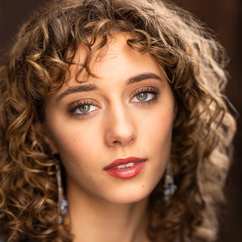 A young woman, Jenna Leigh Miller, with light skin, green eyes, and curly light brown hair gazes at the camera. She wears subtle makeup with rosy lips and dangling earrings. The softly blurred background highlights her serene expression and natural beauty.