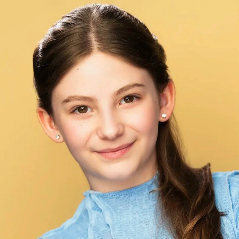 A young girl with fair skin and straight brown hair, partly pulled back, smiles gently at the camera. She wears small stud earrings and a light blue lace top. The solid yellow background gives this Jake Hamilton portrait a warm, cheerful feel.