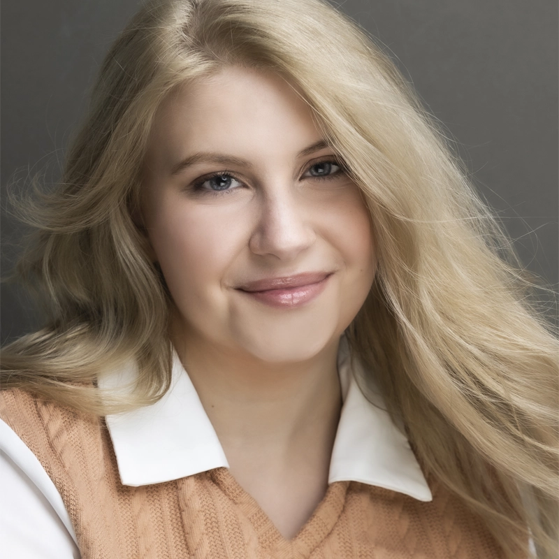 A young woman with long, blonde hair smiles gently. She wears a white collared shirt under a light brown, cable-knit sweater vest. Her hair is parted to the side and flows over her shoulder. Charley Bazz stands against a neutral gray background.