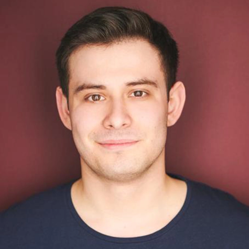 Chris Allyah, a young man with short dark hair and fair skin, smiles softly at the camera. He wears a dark blue crew-neck shirt and stands against a maroon background, with even lighting highlighting his facial features clearly.