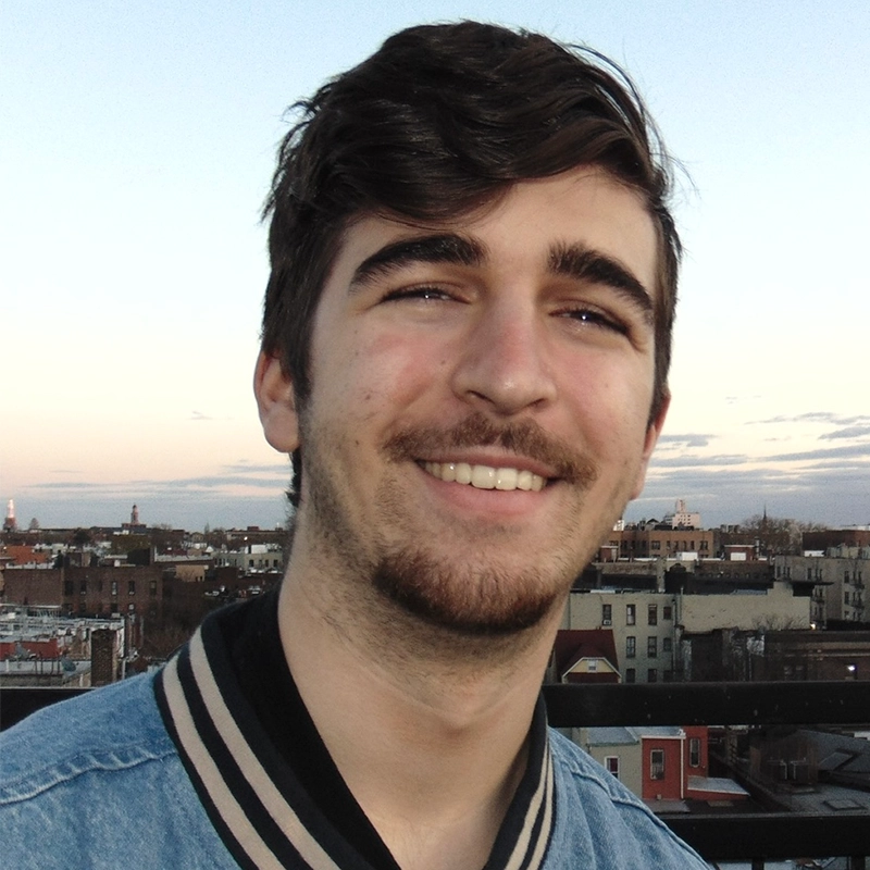 A young man with short brown hair, a light beard, and mustache smiles at the camera. He wears a light blue jacket with black and white collar stripes. Behind him is an urban skyline with buildings and a pale sky at dusk.