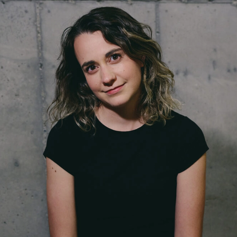 Madeline Benson, with wavy, shoulder-length brown hair and a black short-sleeve top, sits before a gray concrete wall. She gives a gentle smile, tilting her head slightly as she looks directly at the camera.