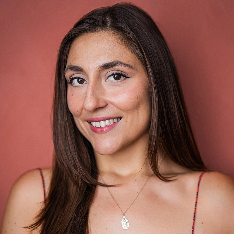 A woman with long brown hair smiles softly in front of a warm reddish-brown background. She wears a thin-strapped top and a gold necklace with an oval pendant. Her makeup is natural, and she looks directly at the camera.