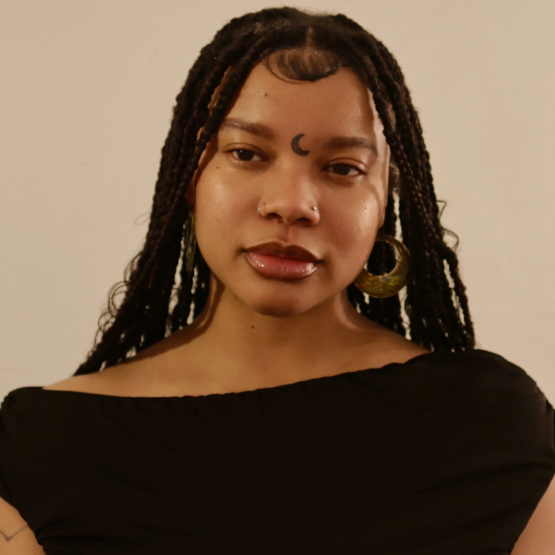 Kamari Branch, a young woman with long braided hair, wears hoop earrings, a nose ring, and a black off-shoulder top. She has a crescent moon symbol on her forehead and looks confidently into the camera against a plain beige background.