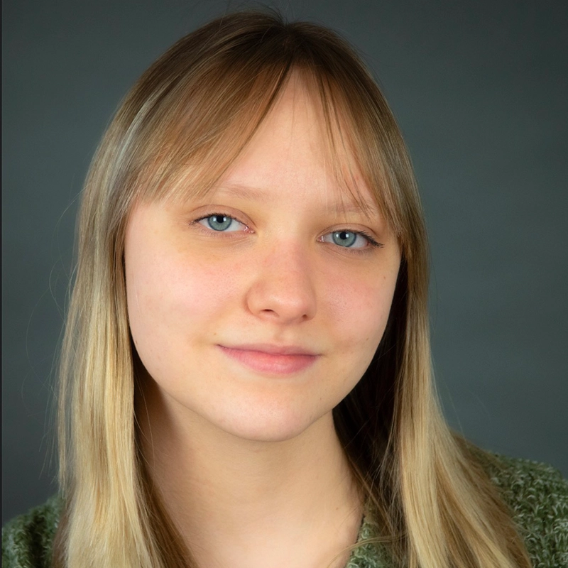 A young woman with long, straight blonde hair and blue eyes is smiling slightly. Gina Marie Incandela is wearing a green sweater and posed against a plain, dark gray background. Her expression is calm and relaxed.